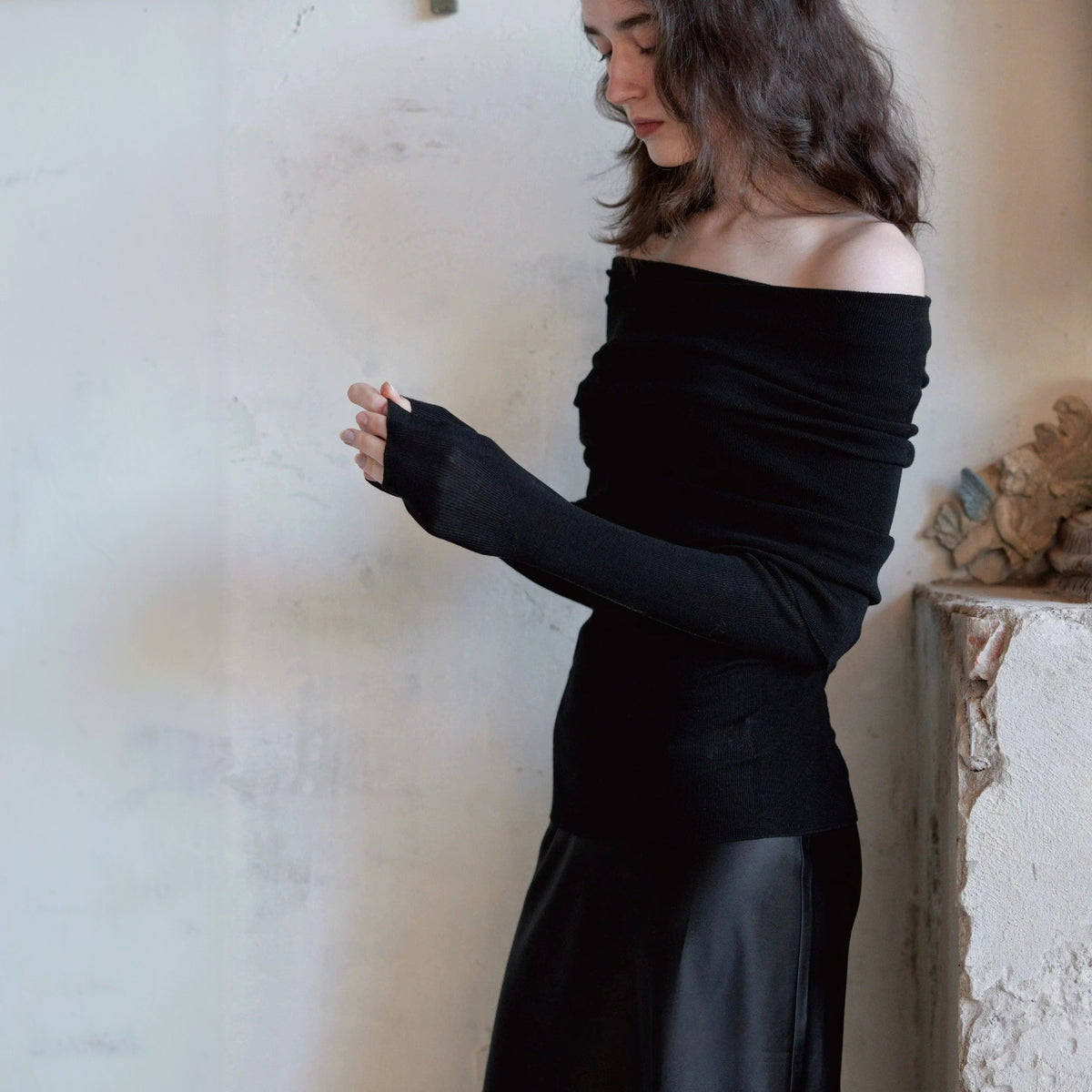 Black off the shoulder sweater with a black skirt, model looking down with one hand at the sleeve cuff
