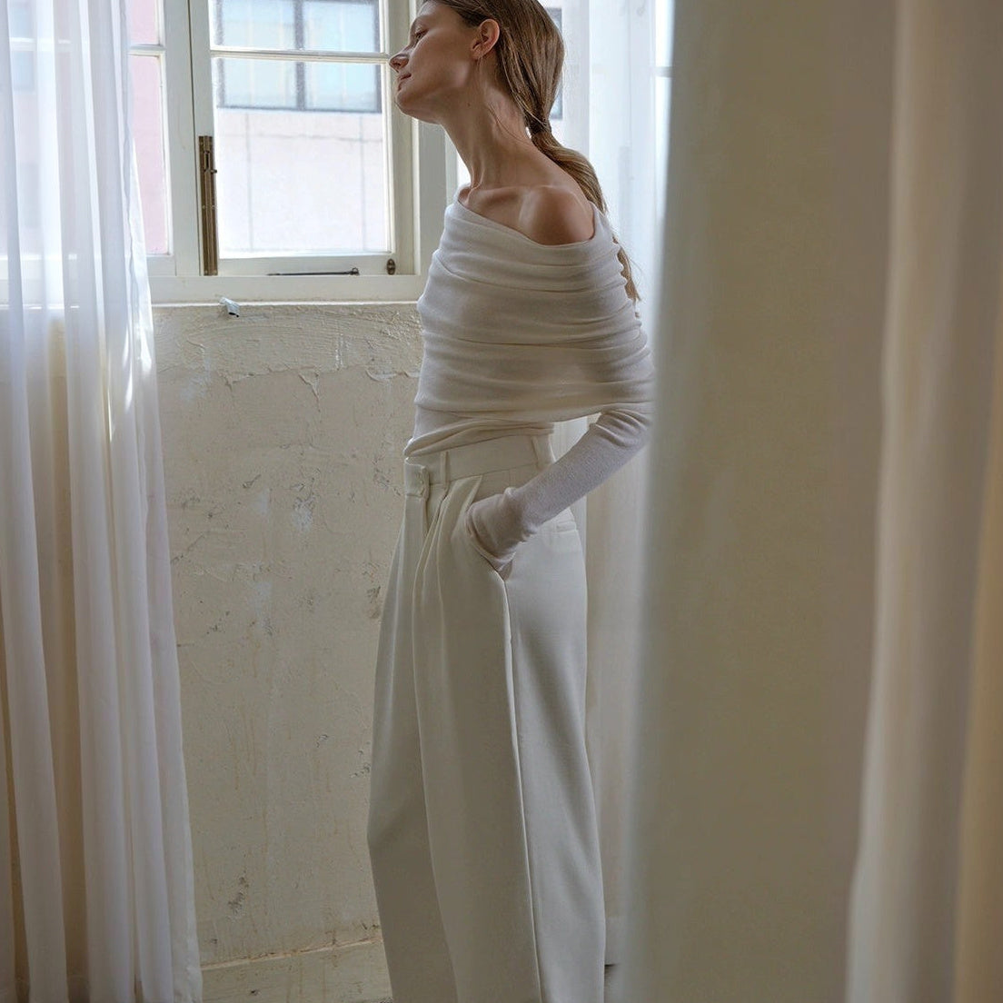 Off the shoulder sweater in white, side view with head tilted back and one hand in the pocket of wide-leg trousers