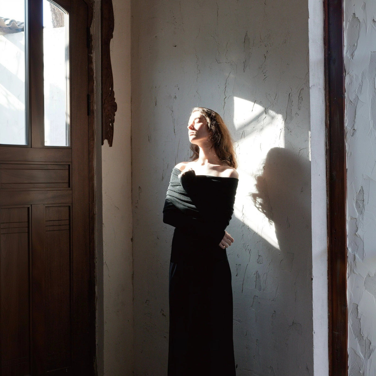 Off shoulder sweater in black with a long black skirt, model in a band of sunlight against a plaster wall