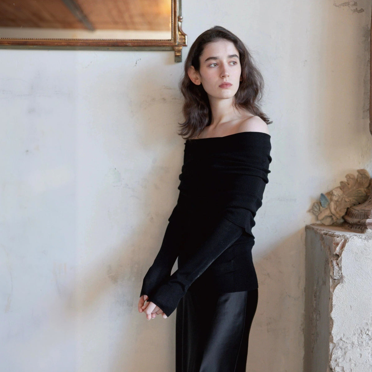 Black off the shoulder sweater and black skirt, model glancing to the side with hands clasped, plaster wall behind