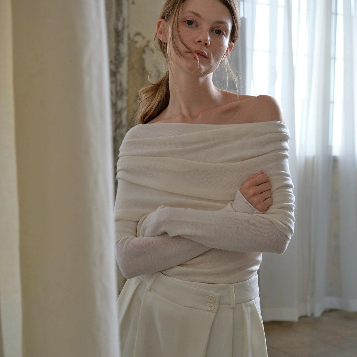 Off the shoulder sweater in white, folded neckline across the shoulders, model with arms crossed in wide-leg trousers
