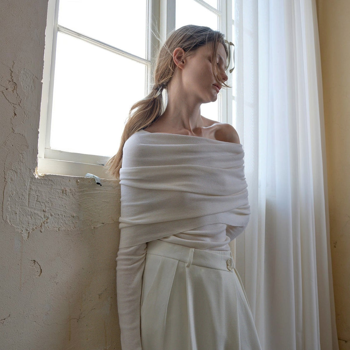 White off the shoulder sweater, three-quarter view at a window with the neckline folded across the bust