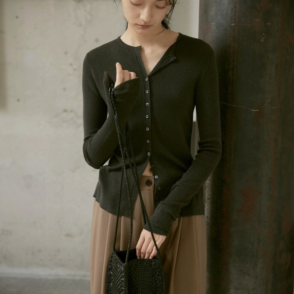 Wool cardigan in dark olive, buttoned, worn with a small woven bag against a concrete wall