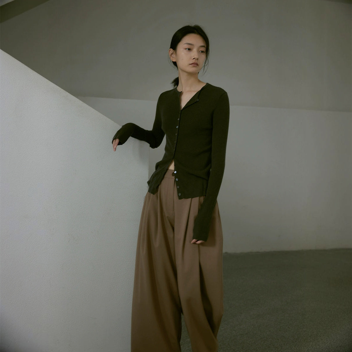 Sheer wool cardigan in dark olive with wide brown trousers, full length beside a stairwell