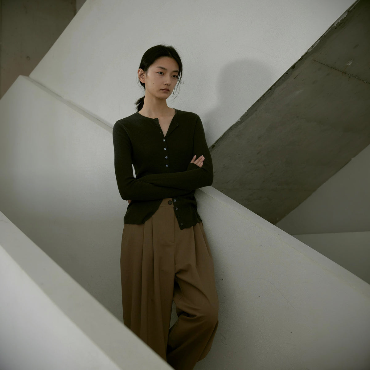 Sheer wool cardigan in dark olive, full length leaning on a stairwell wall with arms crossed