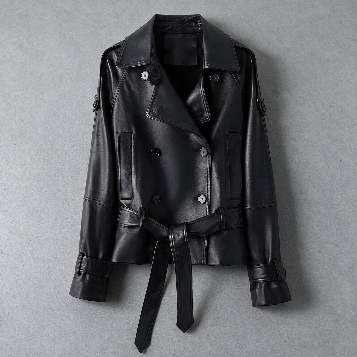 Vegetable-tanned sheepskin leather jacket in black, laid flat with tie belt, on a grey studio backdrop
